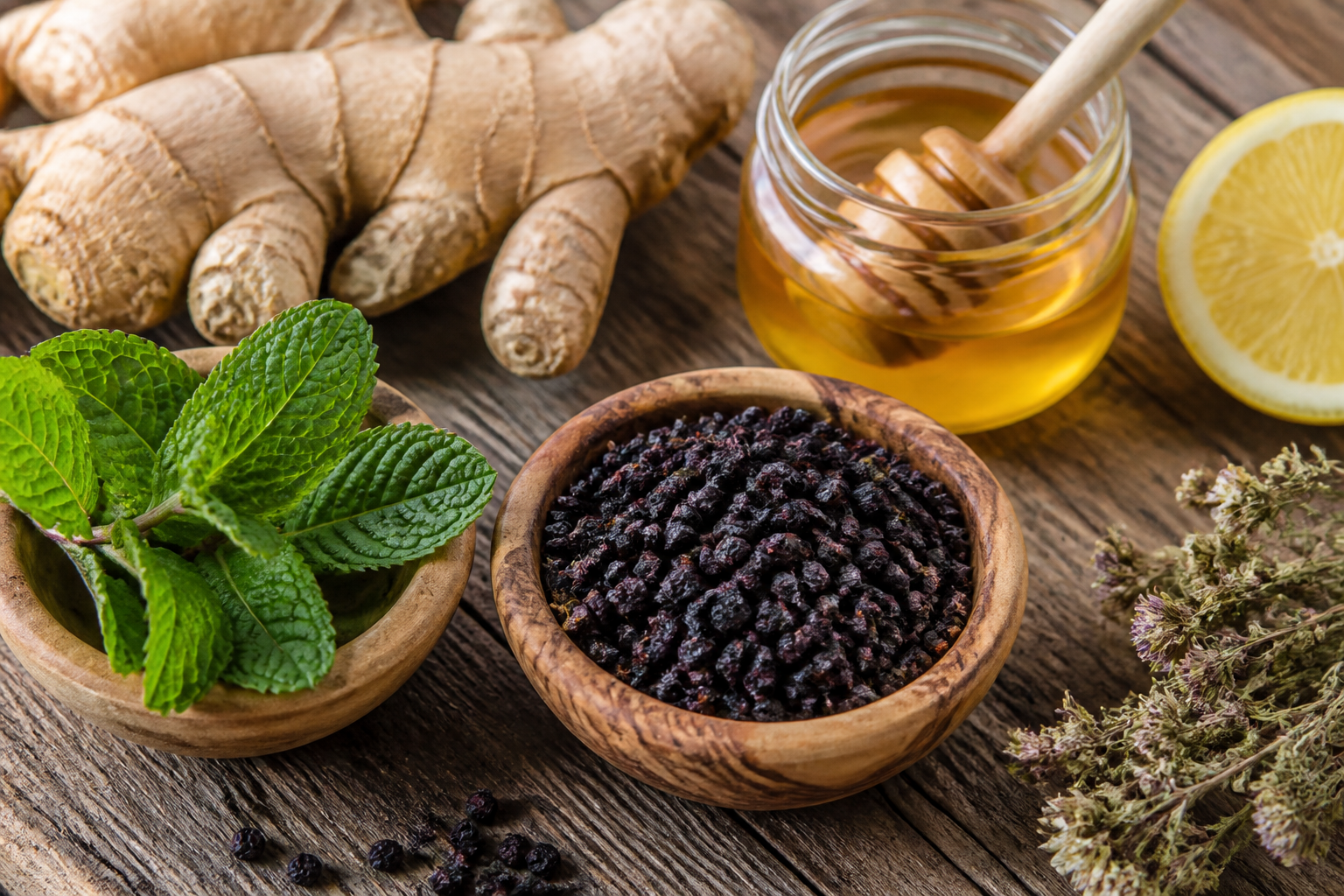 Assorted herbal cold and flu remedies on a wooden surface including raw honey, fresh ginger root, dried elderberries, and peppermint leaves